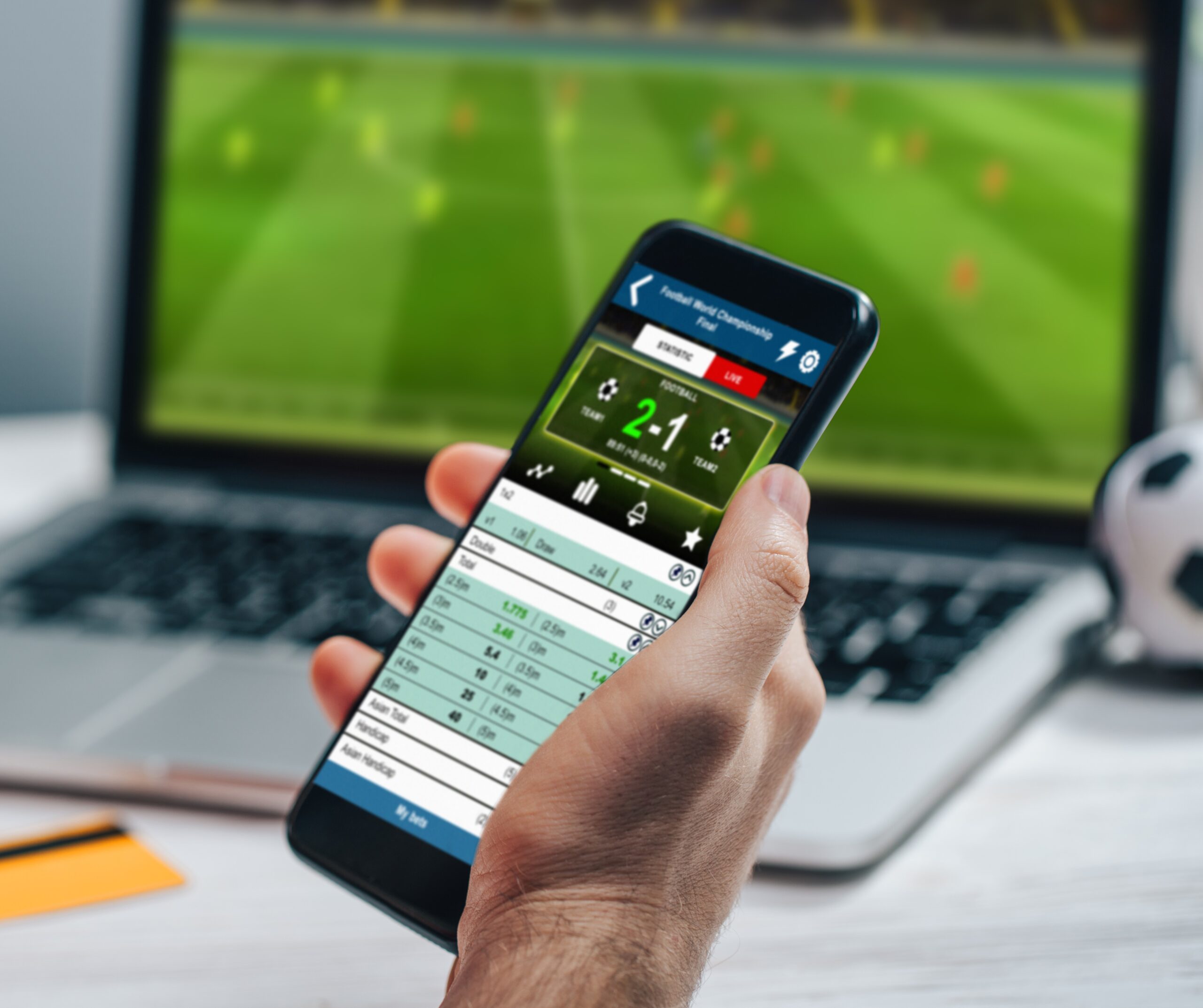 Person holding a phone with a sports betting app on screen and a sports field on a laptop in the background